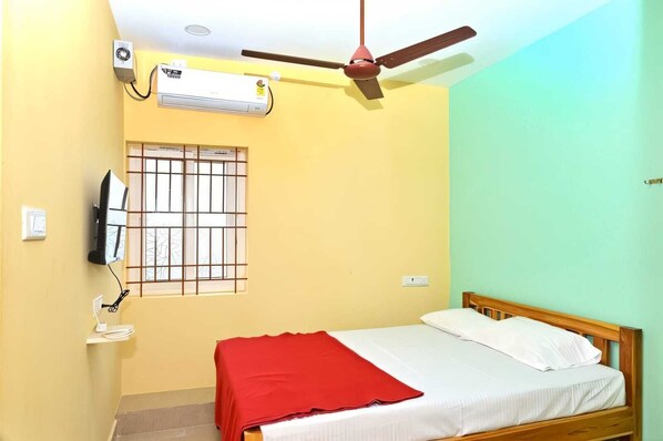A bedroom with a bed, a ceiling fan, an air conditioner, a TV, and a window with bars. - Jothi Mahal (Tiruvannamalai)
