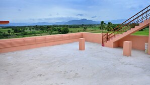 A rooftop terrace with a concrete floor, a staircase with a metal railing, and a view of a green landscape with palm trees and mountains in the distance. - Jothi Mahal (Tiruvannamalai)