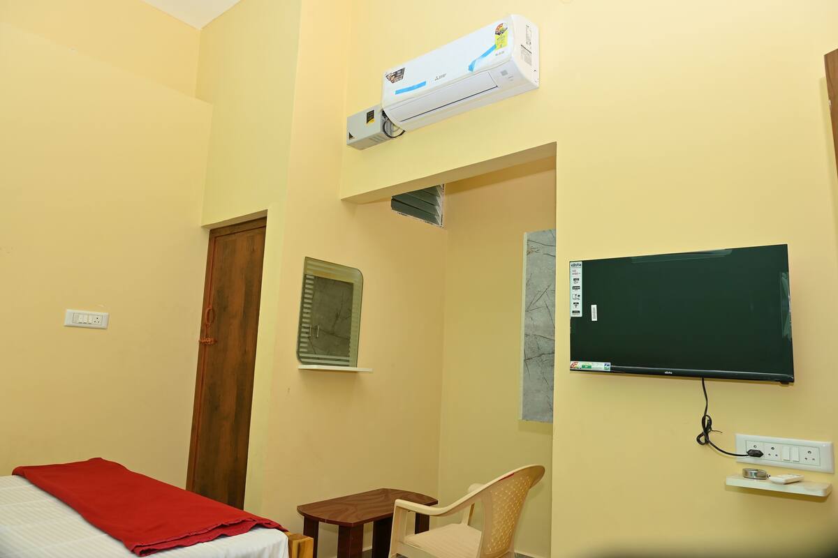 Una habitación con una cama, una colcha roja, una mesita, una silla, un televisor montado en la pared y un aire acondicionado.