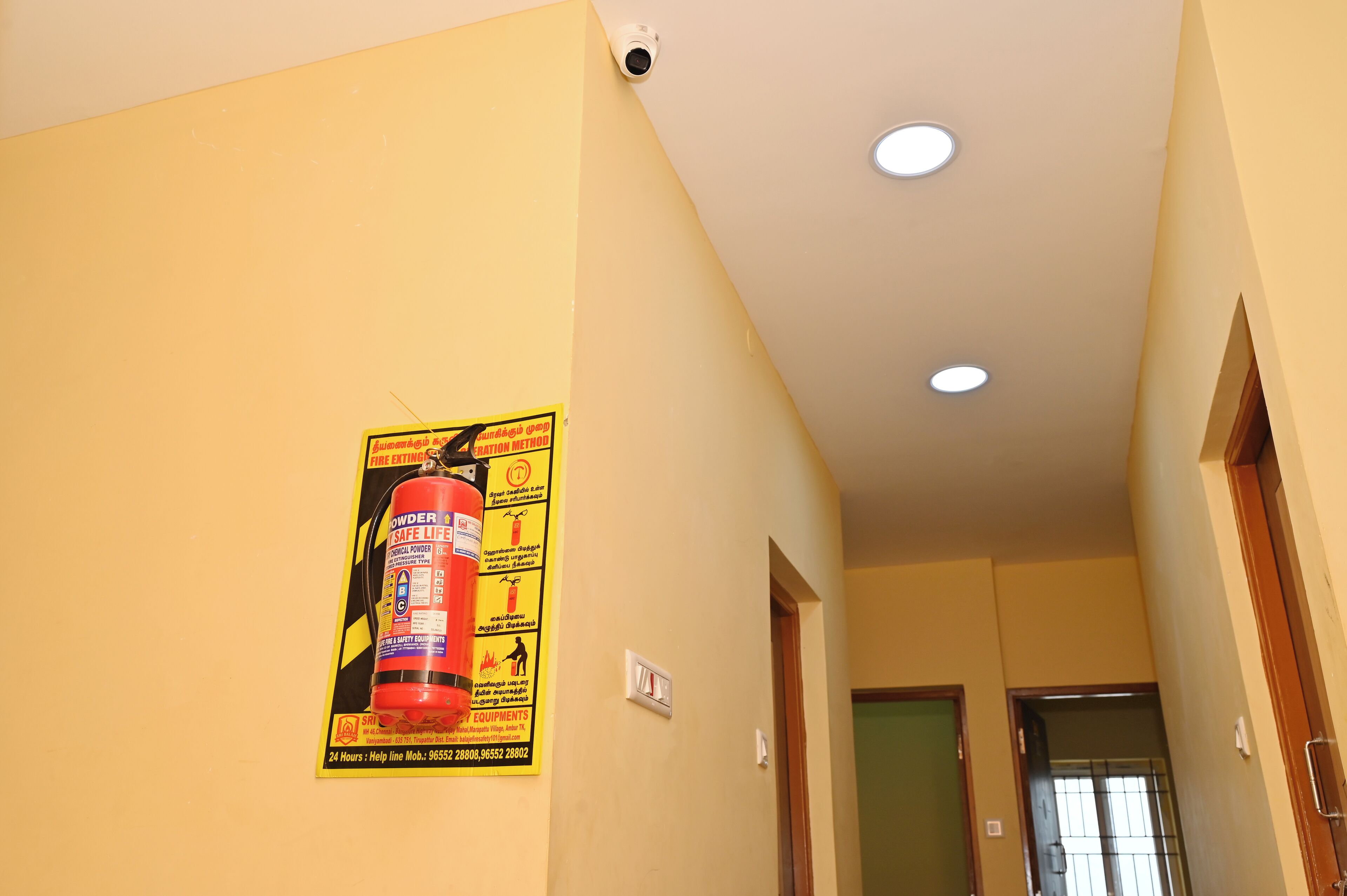 A hallway with a fire extinguisher poster, a security camera, and recessed ceiling lights.