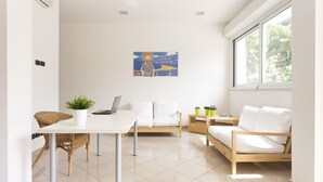 A modern, well-lit room with a white table, two white sofas, and a laptop. There is a large window providing natural light and a view of greenery outside.