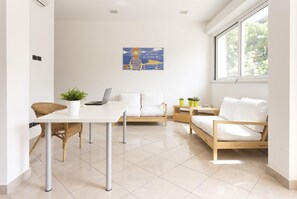 A modern, well-lit room with a white table, two white sofas, and a laptop. There is a large window providing natural light and a view of greenery outside.