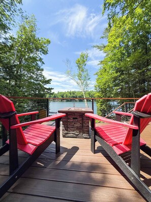 Una terraza con dos sillas Adirondack rojas, un hogar de fuego de piedra y vistas a un lago.
