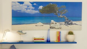 A painting of a beach scene with a tree, rocks, and a clear blue sky. Below the painting, there is a blue shelf with books, a lamp, and a cactus plant.