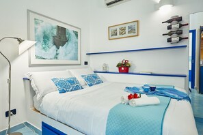 Ein ordentlich eingerichtetes Schlafzimmer mit Bett, Nachttischlampe, einem gerahmten Bild, einer kleinen Ablage mit Flaschen und einer Wand-Klimaanlage.