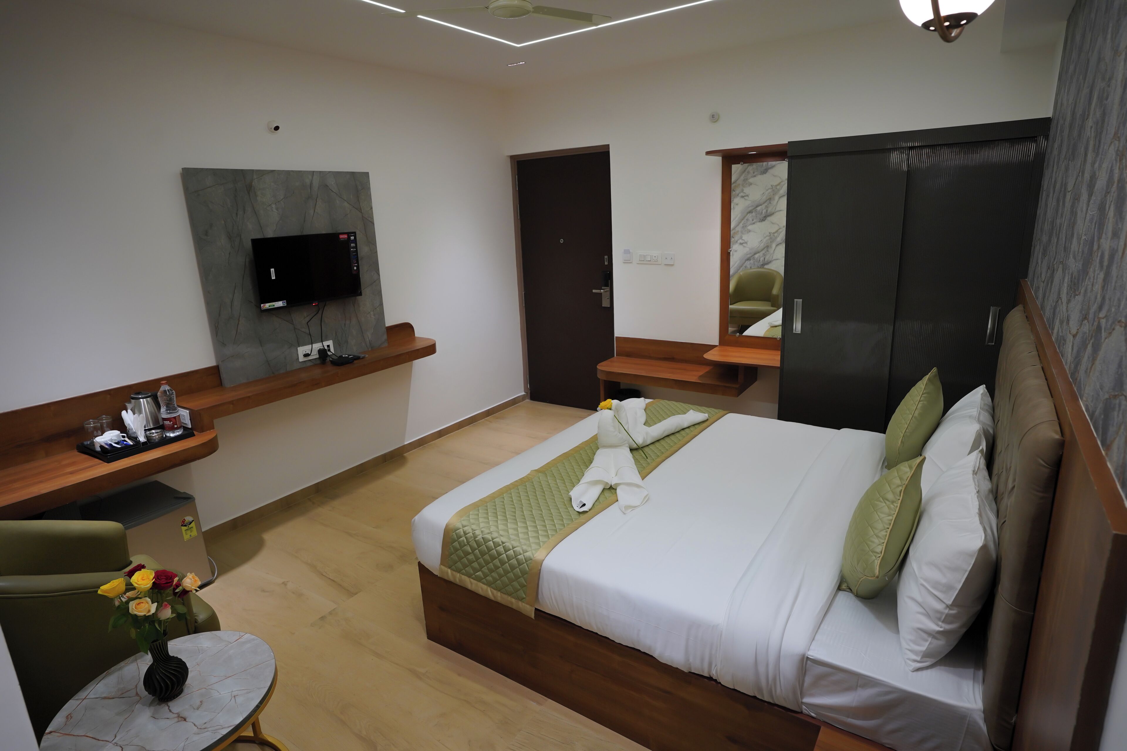 A hotel room with a bed, a TV mounted on the wall, a desk with a chair, and a small table with a vase of flowers.