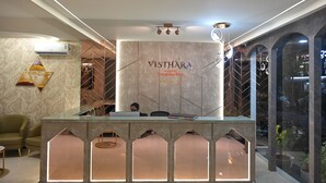 A reception desk with a marble wall and the name "Vishthara" prominently displayed. There are two people seated at the desk, and a potted plant is visible in the background. - VISTHARA a unit of Suraksha Stay (Bangalore)
