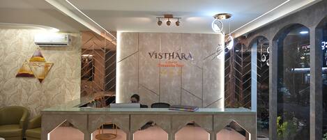 A reception desk with a marble wall and the name "Vishthara" prominently displayed. There are two people seated at the desk, and a potted plant is visible in the background.