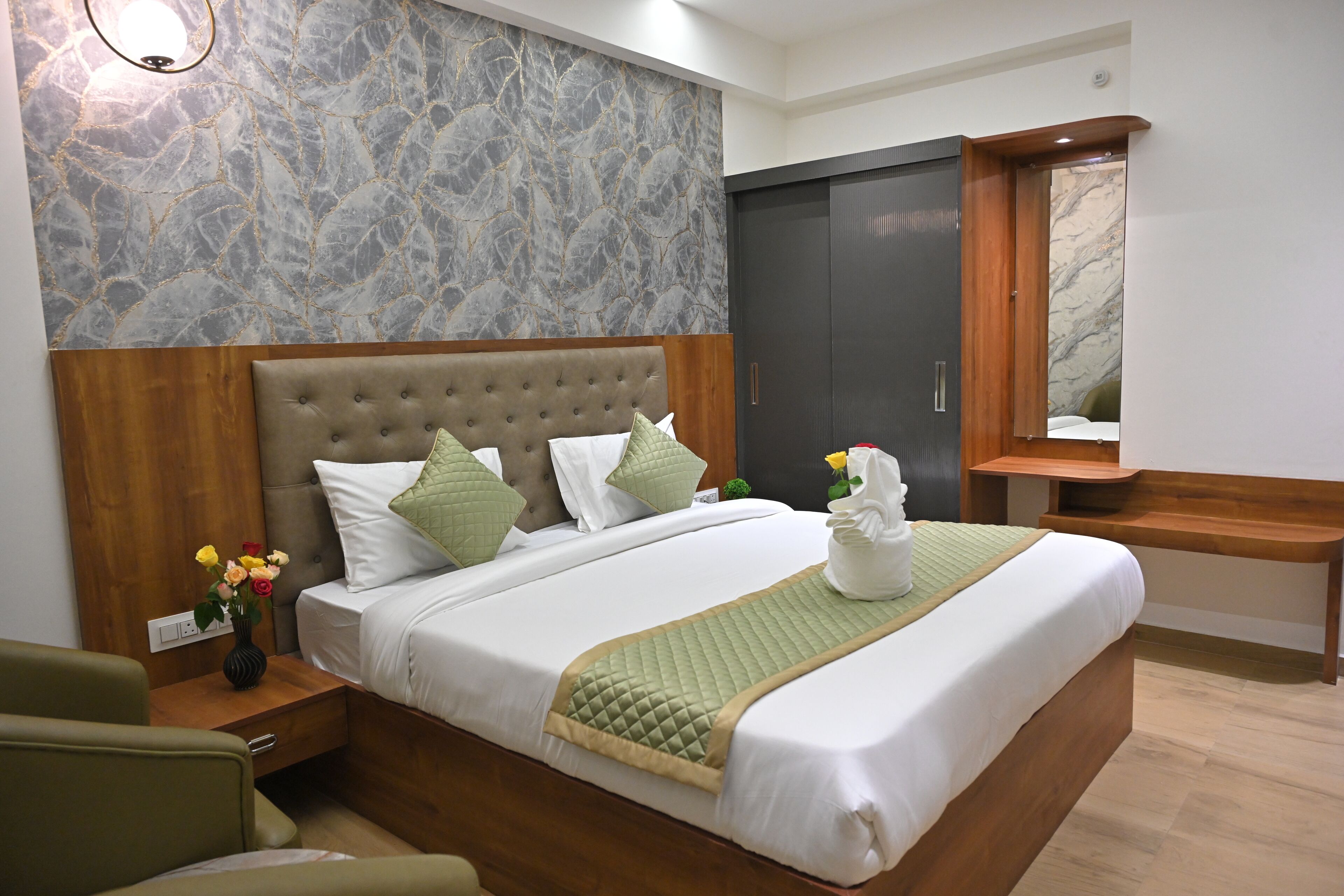 A neatly arranged hotel room with a large bed, a wooden headboard, a marble-patterned wall, a dark wardrobe, and a small bedside table with a vase of flowers.