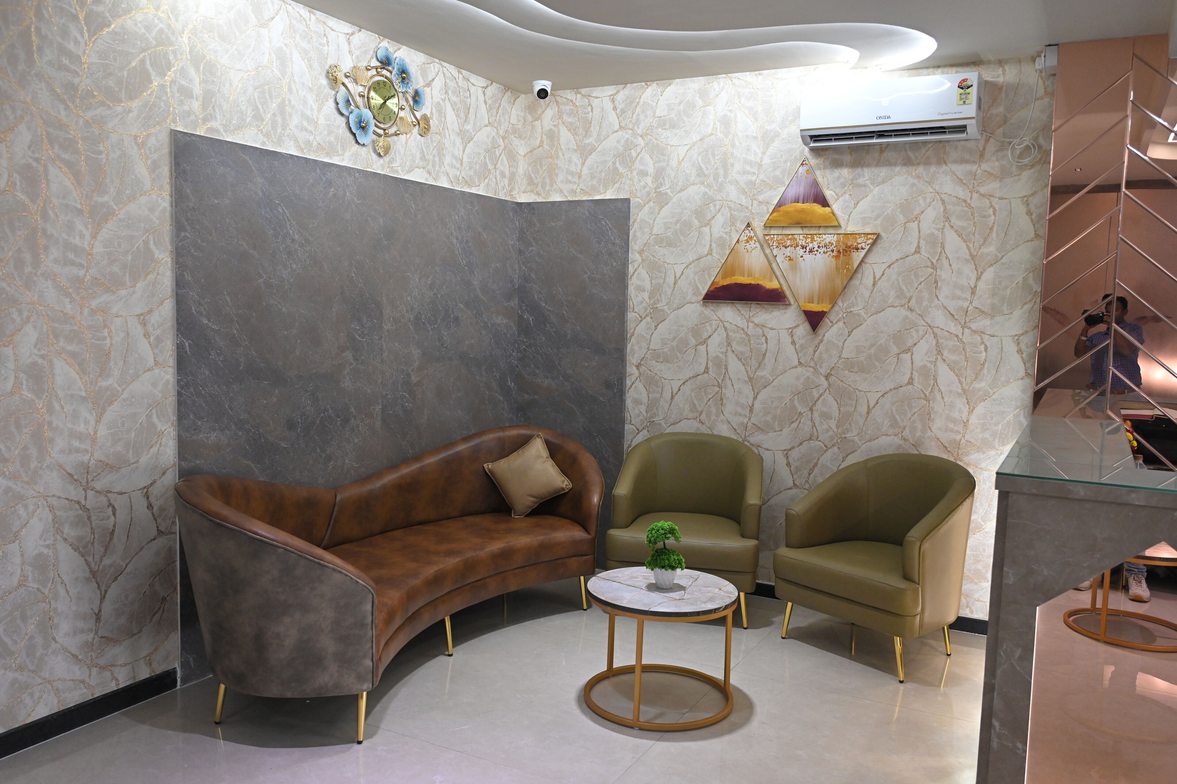 Un lobby moderno con un sofá de cuero marrón curvo, dos sillones verdes, una pequeña mesa redonda con una planta y una pared con estampado de mármol y una pieza decorativa.