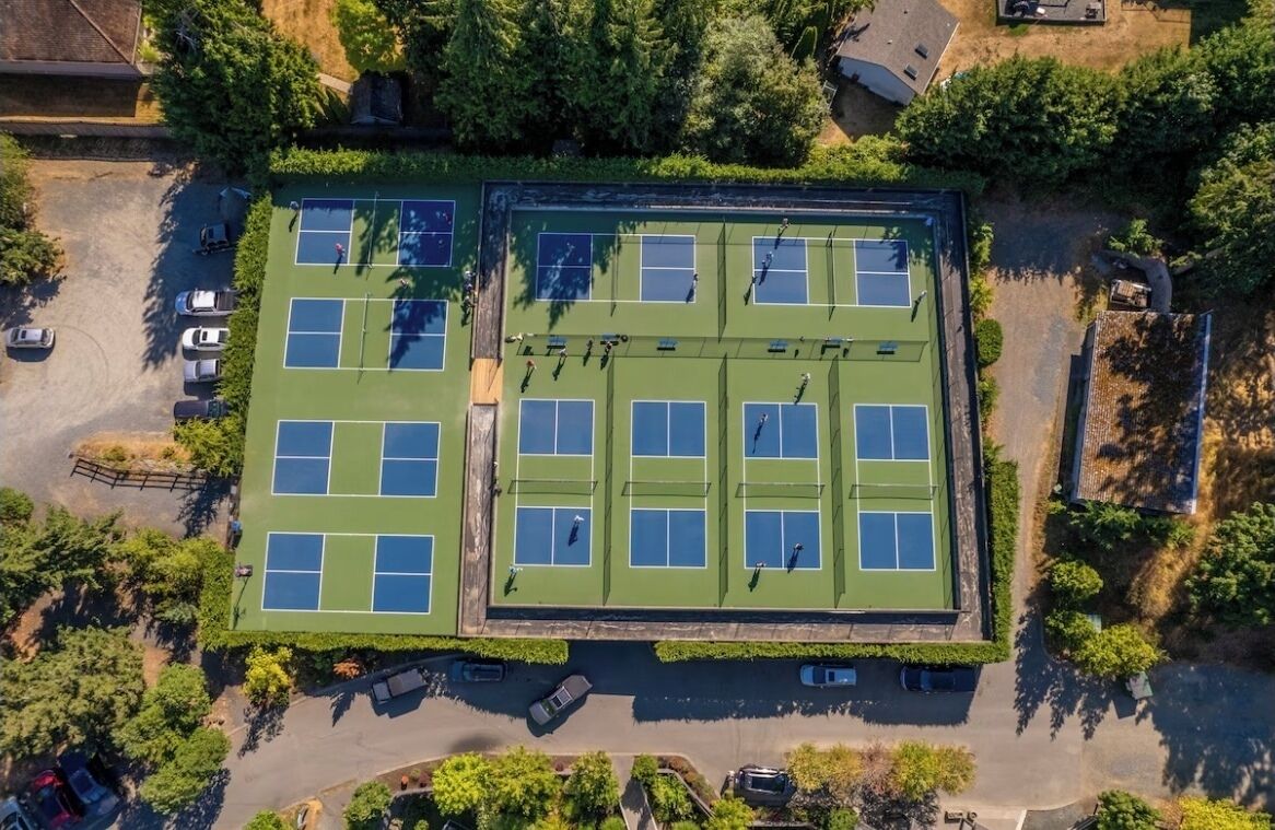 Vue aérienne d’un complexe de courts de tennis comprenant plusieurs terrains, entouré d’arbres et bordé de zones de stationnement.