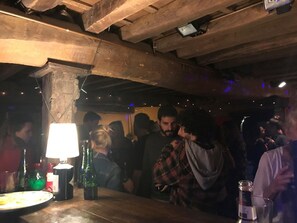 Eine belebte Bar mit Holzbalken, einem Tisch mit Flaschen und einer Person in einem Karohemd.
