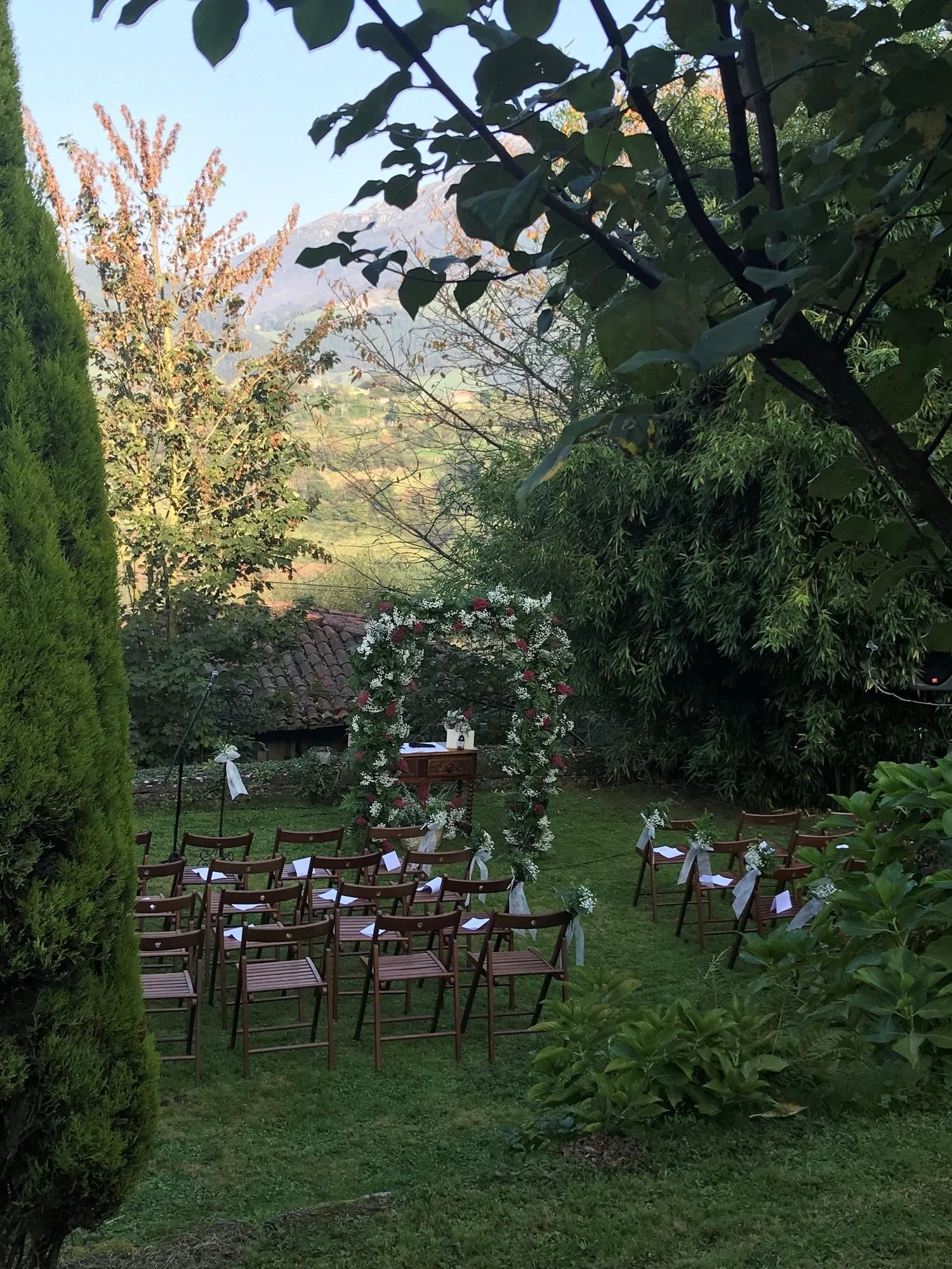 Un lieu de réception en plein air pour les mariages, avec des chaises en bois disposées en rangées, une arche décorée de fleurs et un décor composé d’arbres et de collines.