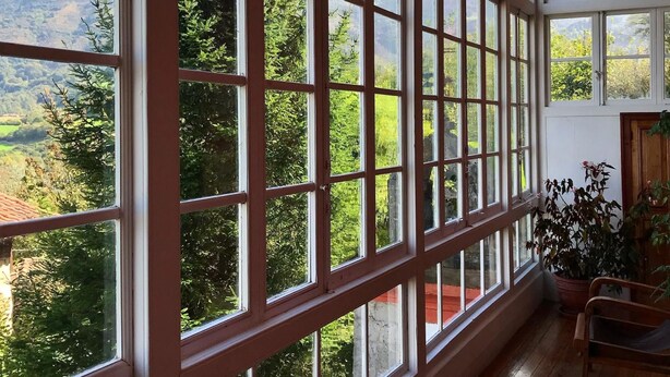 A room with large windows offering a view of a mountainous landscape. There are wooden chairs and a potted plant inside. - Hotel Palacio De Libardon (Colunga)