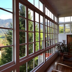 Ein Zimmer mit großen Fenstern und Blick auf eine gebirgige Landschaft. Holzstühle und ein Blumentopf befinden sich im Raum.