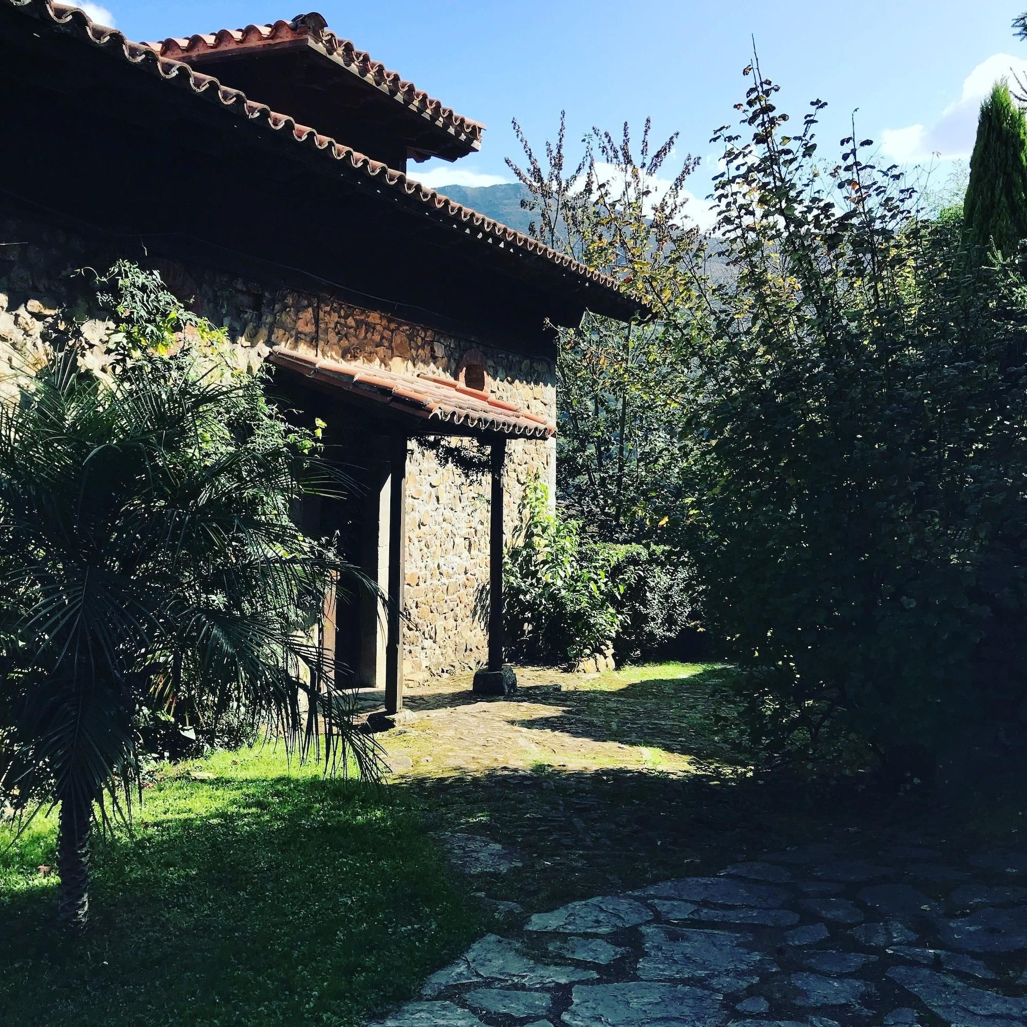 A rustic stone building with a tiled roof, surrounded by lush greenery and a cobblestone path.
