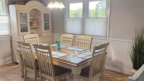 Un comedor con una mesa y sillas de madera, un armario blanco con puertas de cristal y una ventana con persianas.