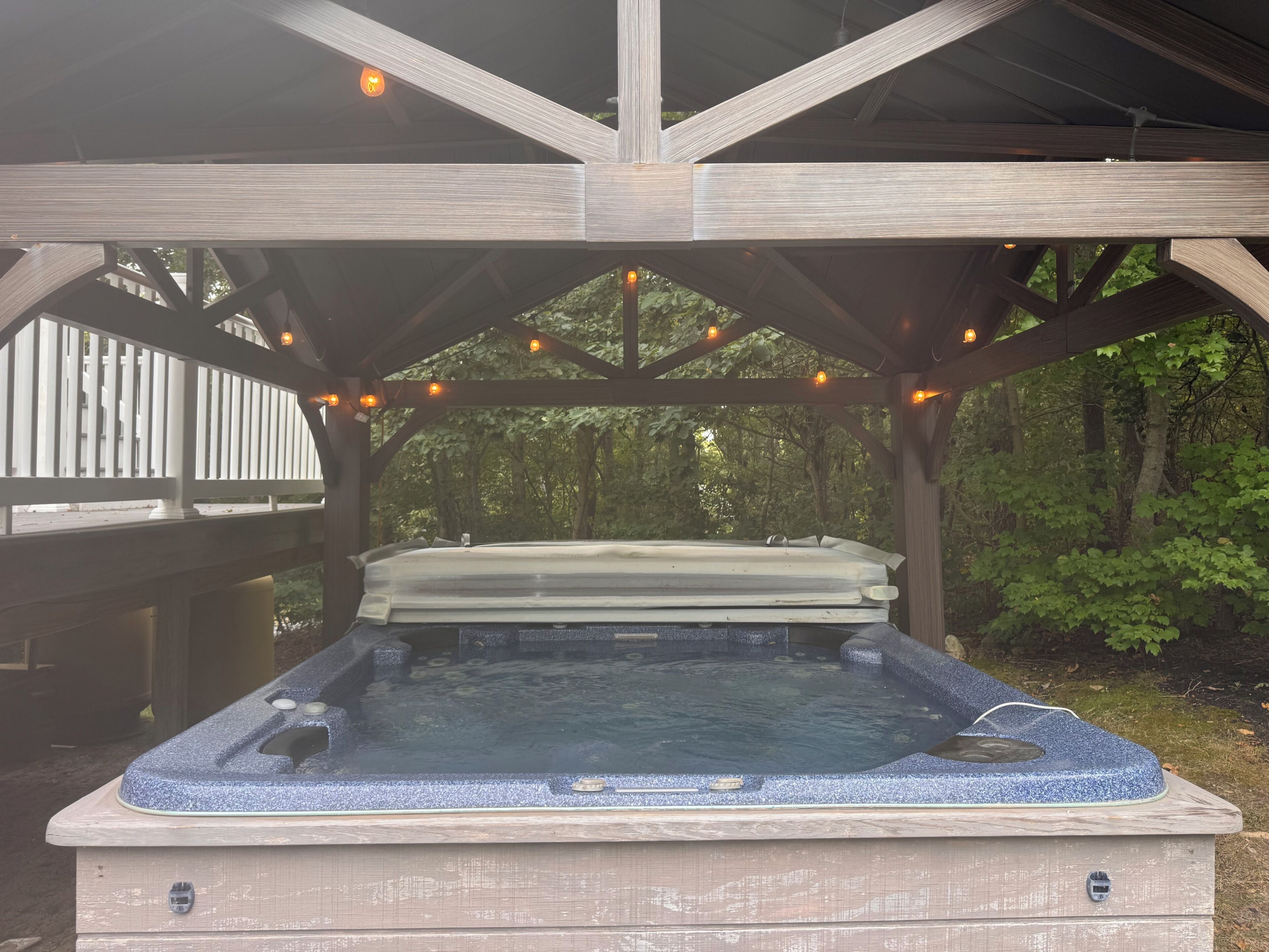 A hot tub under a wooden pergola with string lights, surrounded by trees.