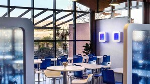 A modern indoor dining area with round tables and blue chairs, large windows, and a brick wall. - Ibis Budget Madrid Alcala de Henares (Alcala de Henares)