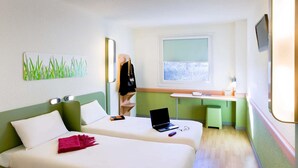 A hotel room with two beds, a desk with a laptop, a window with a view, and a coat rack with a jacket. - Ibis Budget Madrid Alcala de Henares (Alcala de Henares)