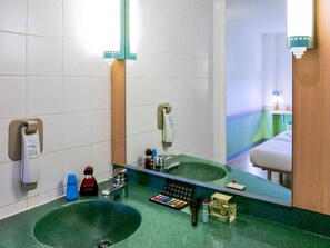 Un baño con un lavabo verde, un espejo, un secador de manos, dispensador de jabón y un estante pequeño con artículos de tocador.