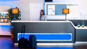 A hotel reception area with a reception desk, a computer monitor, and a sign that reads "RECEPCIÓN". There are two potted plants, a blue illuminated panel, and a blue wall. - Ibis Budget Madrid Alcala de Henares (Alcala de Henares)