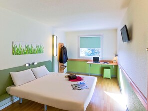 Habitación de hotel con cama, escritorio, silla, televisor y ventana con vista a árboles.