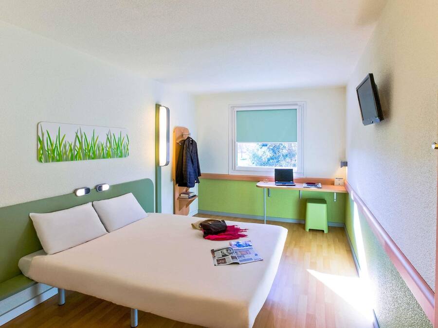 A hotel room with a bed, a desk with a laptop, a TV, and a window with a view of trees.