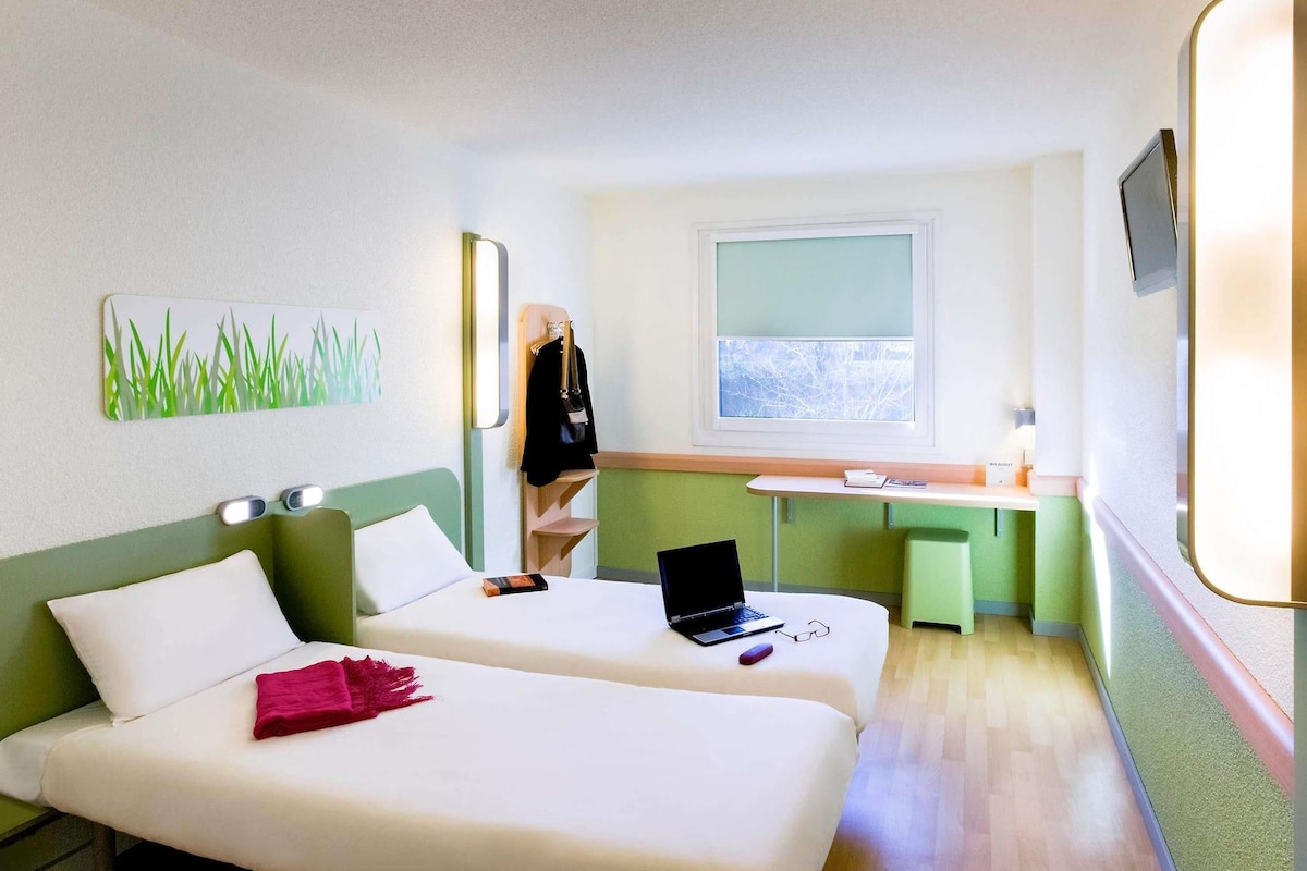 A hotel room with two beds, a desk, a laptop, and a window with a view.