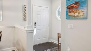 A hallway with a white door, a doormat, a wooden chair, and a painting of a boat.