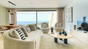 A modern living room with a large sofa, armchairs, a coffee table, and a TV. There is a large window with a view of the ocean.