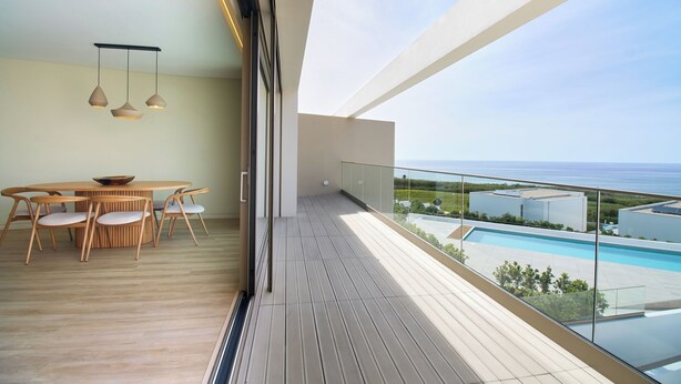 A modern balcony with a wooden floor, a round dining table with chairs, and a view of a swimming pool and buildings.