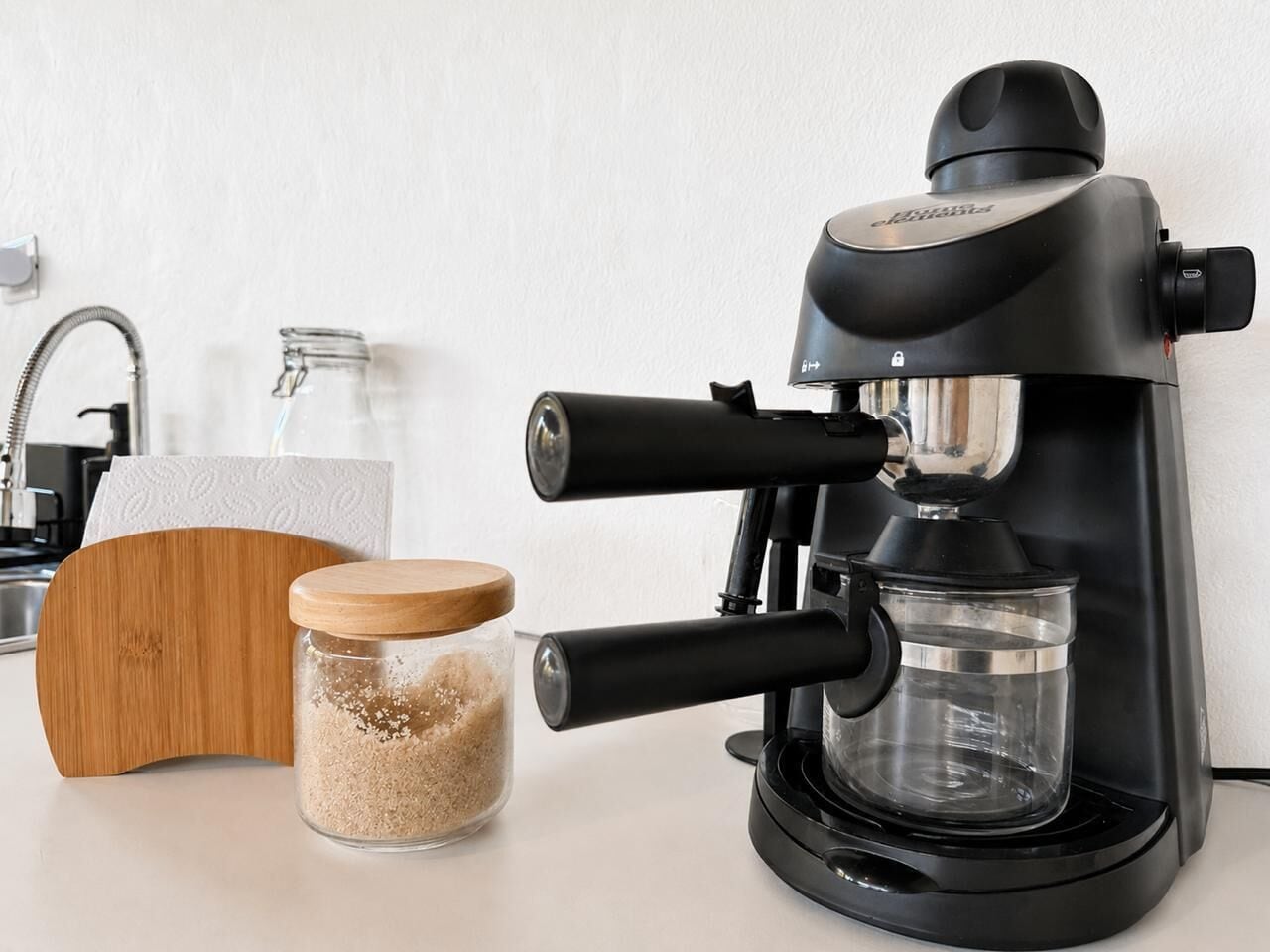 A coffee maker with a water reservoir, a glass jar with a lid, and a wooden cutting board.