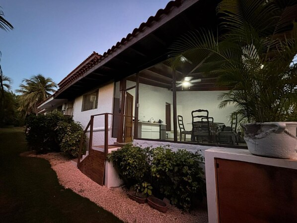 Una casa de dos pisos con techo de tejas, un patio con mesa y sillas, y una planta grande en maceta.