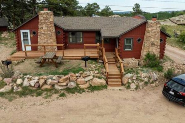 A rustic cabin with a stone chimney, wooden deck, and a dirt driveway.