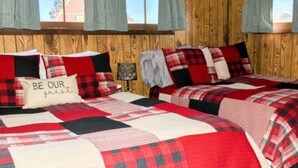 Quarto com duas camas, roupa de cama xadrez, almofada com a inscrição 'BE OUR guest', paredes de madeira e janela com cortinas.
