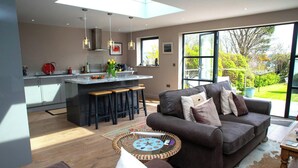 Eine moderne Küche mit Kochinsel, eine Essecke mit Sofa und Couchtisch sowie große Fenster mit Blick ins Freie.