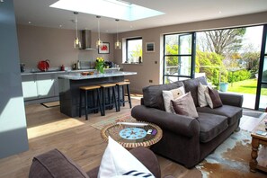 A modern kitchen with a central island, a dining area with a sofa and a coffee table, and large windows offering a view of the outdoors. - Green Meadows, Newport (Newport)