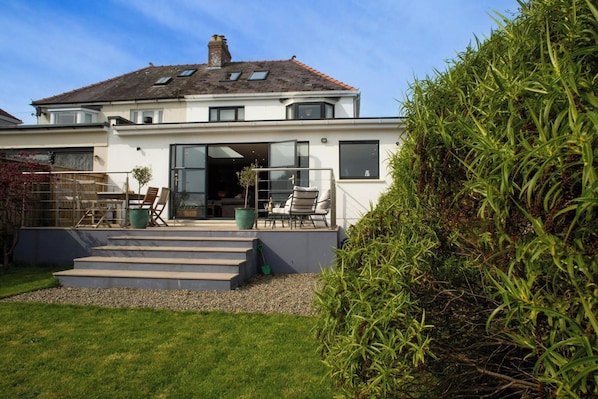 A modern two-story house with a large glass door entrance, a patio with outdoor furniture, and a well-maintained garden.