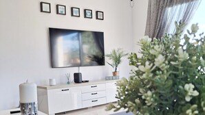 Una sala moderna con un televisor de pantalla plana montado en la pared, una consola blanca con una bandeja decorativa, una planta en maceta y una cortina gris.