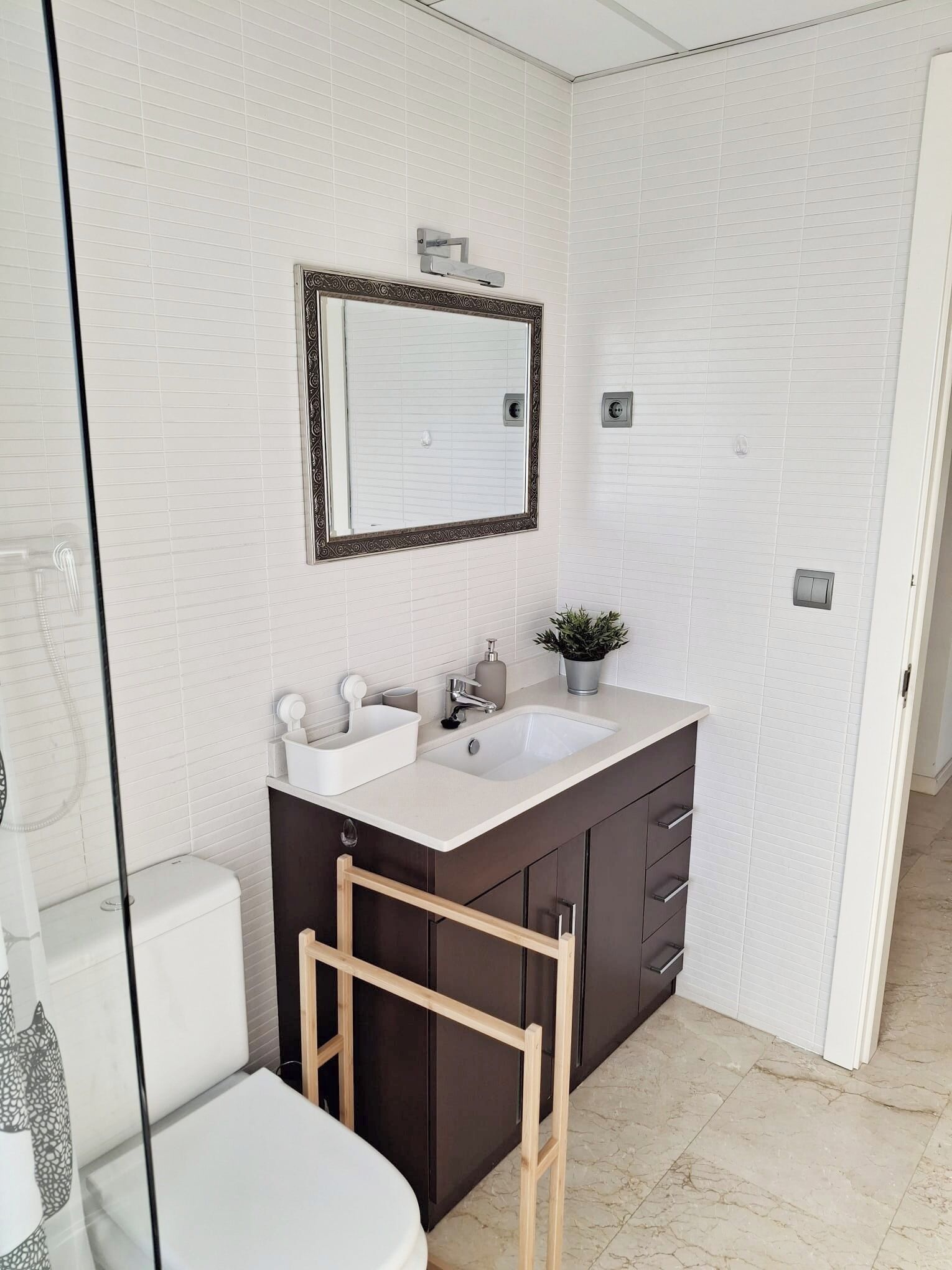 A bathroom with a white tiled wall, a large mirror, a double sink vanity, a small potted plant, and a wooden ladder.