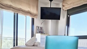 Un dormitorio con una cama, un televisor montado en la pared y vistas al exterior a través de amplias ventanas.