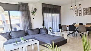 Una sala de estar moderna con un sofá seccional gris, mesas de centro blancas y un comedor con una mesa y sillas de madera.