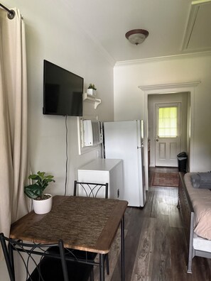 A compact living space with a dining table, chairs, a flat-screen TV, a small refrigerator, a potted plant, and a sofa.