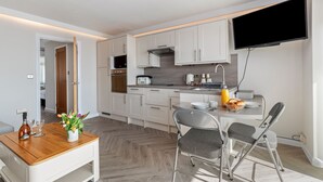 A modern kitchen with white cabinets, a flat-screen TV mounted above the counter, and a dining area with a wooden table and metal chairs. - 9 Strandways - Sea Views Parking Beach Access (Saundersfoot)