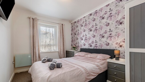 A bedroom with a bed, nightstands, a dresser, a radiator, and a window with curtains. - Little Haven - Cosy Tenby Apartment (Tenby)