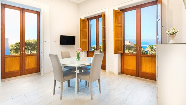 Ein Essbereich mit einem runden Tisch, vier Stühlen und einer Vase mit Blumen. Zwei große Fenster mit Holzrahmen bieten Blick auf das Meer.