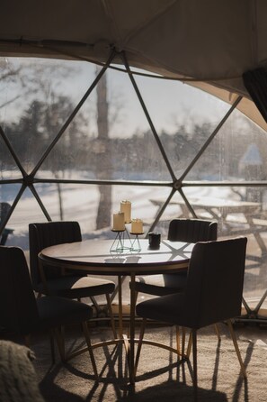 Une table ronde entourée de quatre chaises, agrémentée de bougies et d’une tasse, le tout installé dans une structure ressemblant à une tente, d’où l’on peut admirer les arbres et la neige à l’extérieur.