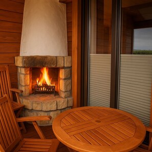 A stone fireplace with a fire, wooden chairs, and a round table in a cozy outdoor setting.