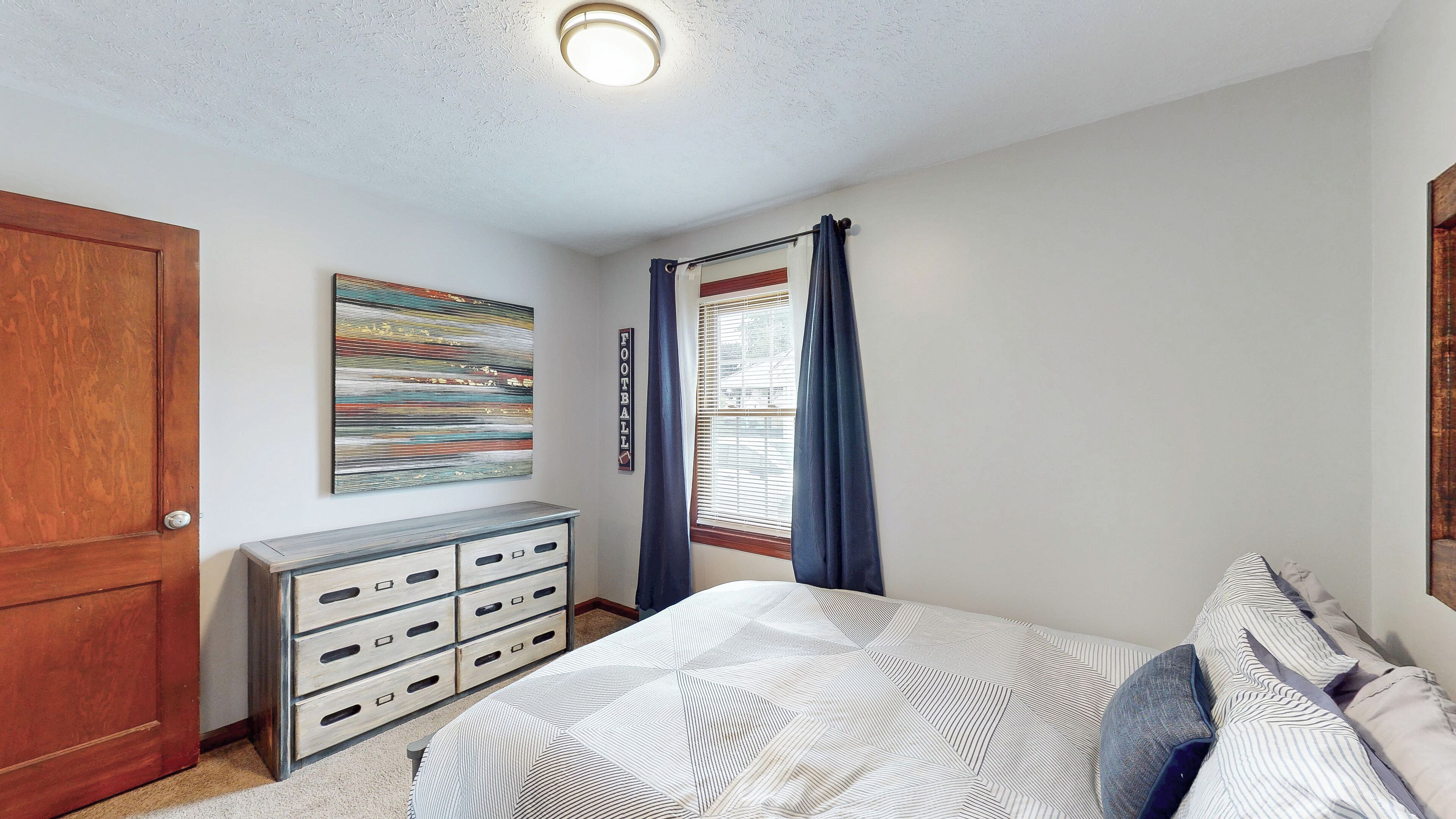 A bedroom with a bed, a dresser, a painting, and a window with curtains.